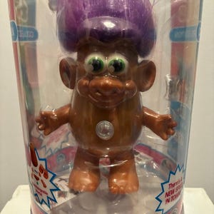 Puede incluir: Muñeca Good Luck Trolls en un embalaje de plástico transparente. La muñeca tiene piel marrón, ojos verdes grandes y pelo morado y puntiagudo. La muñeca está de pie con los brazos extendidos. El embalaje tiene las palabras "Good Luck Trolls" en rojo y blanco.