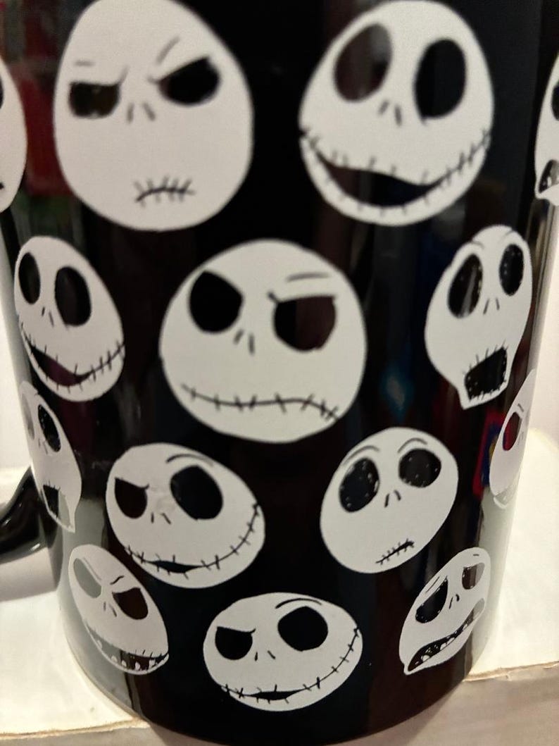Puede incluir: Taza de caf&eacute; negra con caras blancas ovaladas con sonrisas cosidas y diversas expresiones. El dise&ntilde;o de la taza est&aacute; inspirado en una popular pel&iacute;cula de animaci&oacute;n, con un patr&oacute;n repetido de la cara del personaje.