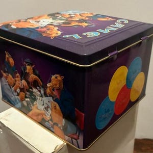 May include: A purple metal box with a hinged lid. The box features illustrations of a camel playing poker and poker chips. The word "CAMEL" is printed on the lid in teal letters. The box has gold trim.