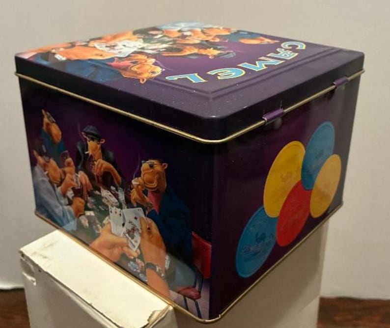 May include: A purple metal box with a hinged lid. The box features illustrations of a camel playing poker and poker chips. The word "CAMEL" is printed on the lid in teal letters. The box has gold trim.