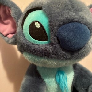 May include: A plush toy character with soft blue fur, large ears with pink inner lining, and a dark blue nose. The character features bright turquoise accents around the eyes and on the chest, with a small tuft of blue hair on top of the head.