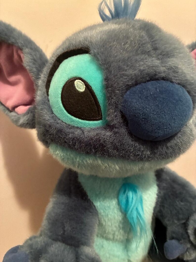 May include: A plush toy character with soft blue fur, large ears with pink inner lining, and a dark blue nose. The character features bright turquoise accents around the eyes and on the chest, with a small tuft of blue hair on top of the head.