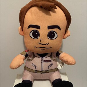 May include: A plush doll of a character from Ghostbusters, featuring a light pink jumpsuit with the name "VENKMAN" on the chest. The doll has brown hair, large eyes, and black shoes. The overall design is cartoonish.