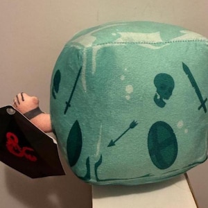 May include: A plush, cube-shaped toy in shades of teal, featuring illustrations of a skull, swords, a shield, and an arrow. The toy is paired with a black shield-shaped tag with red text. The toy is designed for role-playing games.