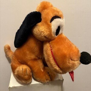 May include: A vintage plush toy dog with orange fur, black ears, and a black nose. The dog has a red tongue sticking out and large white eyes with black pupils. The toy is sitting on a white surface.