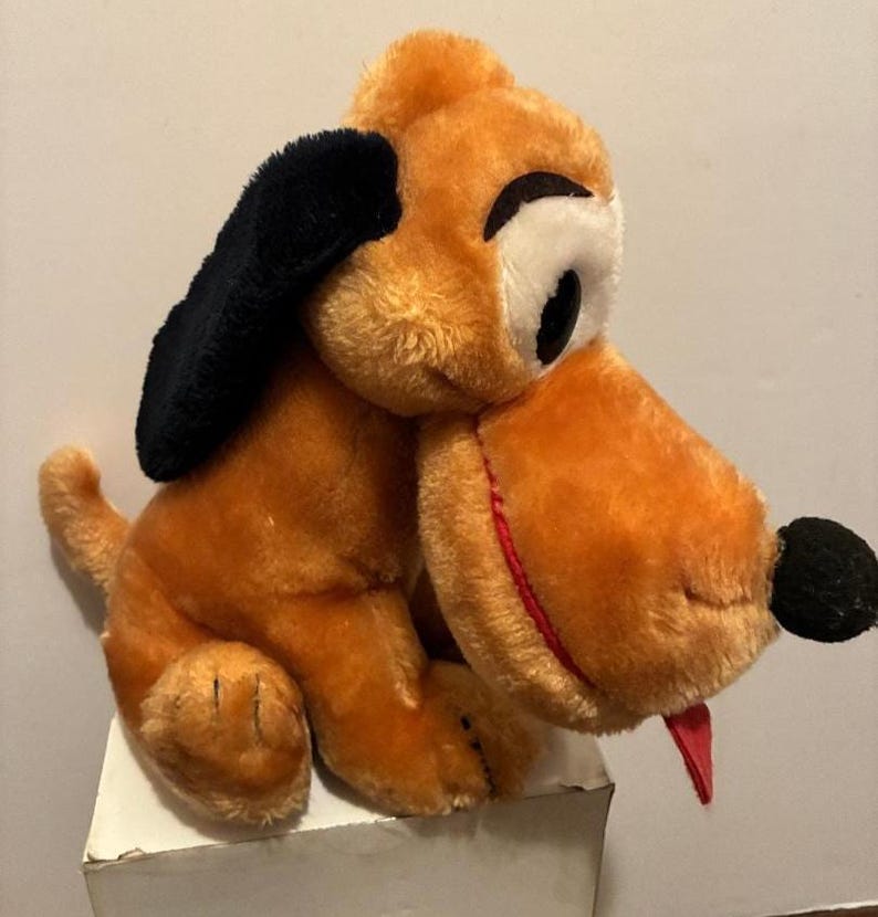 May include: A vintage plush toy dog with orange fur, black ears, and a black nose. The dog has a red tongue sticking out and large white eyes with black pupils. The toy is sitting on a white surface.