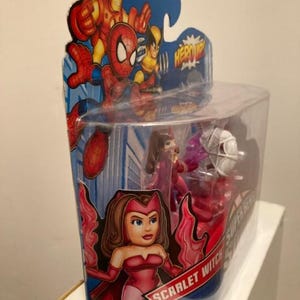 May include: A packaged Scarlet Witch action figure from the Super Hero Squad series. The figure is in a clear plastic container with a cardboard backing featuring Iron Man, Spider-Man, and Wolverine. The Scarlet Witch figure is red and pink.