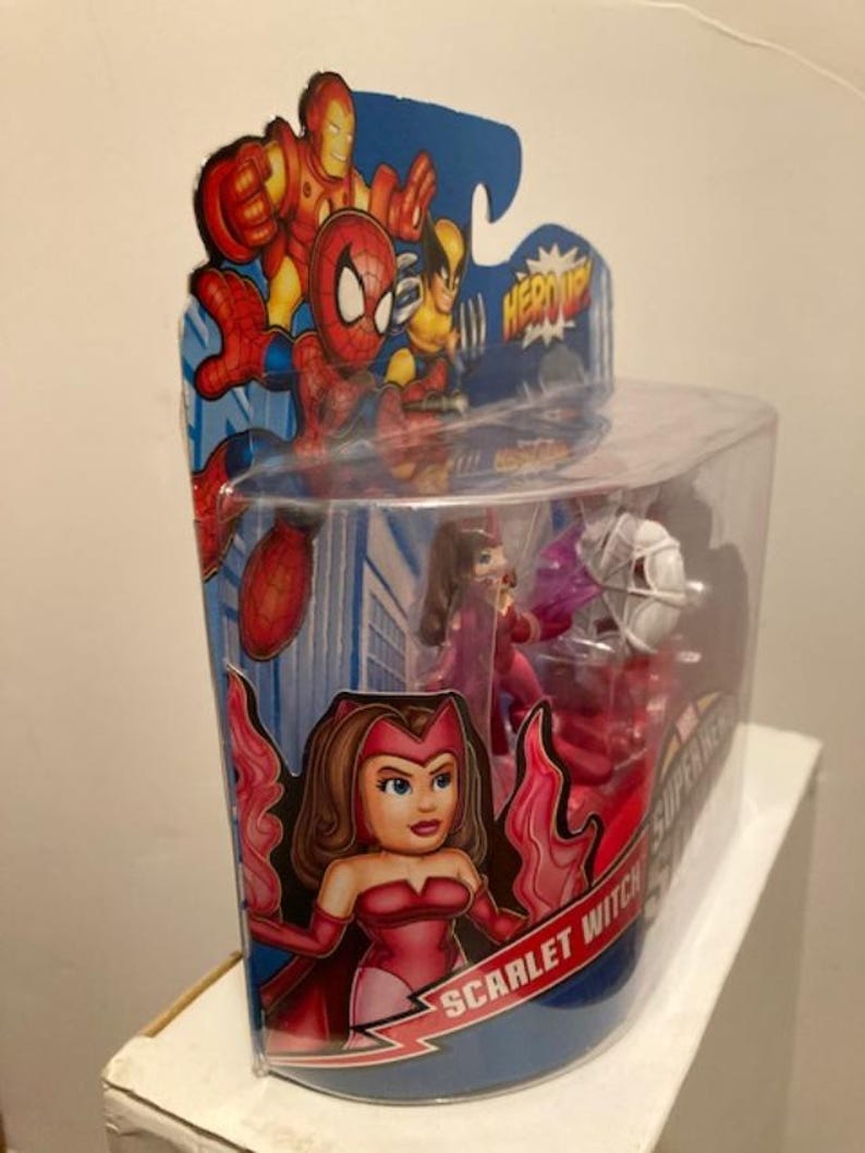 May include: A packaged Scarlet Witch action figure from the Super Hero Squad series. The figure is in a clear plastic container with a cardboard backing featuring Iron Man, Spider-Man, and Wolverine. The Scarlet Witch figure is red and pink.