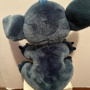 May include: A plush toy of the character Stitch, featuring dark blue fur, large ears, and a small tuft of light blue hair on top of the head. The figure is shown from the back, displaying a dark blue patch along the spine and dark blue claws.