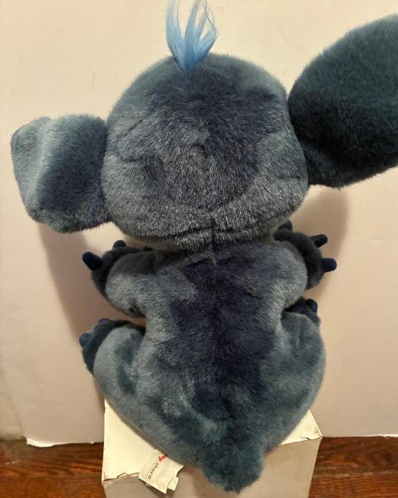 May include: A plush toy of the character Stitch, featuring dark blue fur, large ears, and a small tuft of light blue hair on top of the head. The figure is shown from the back, displaying a dark blue patch along the spine and dark blue claws.