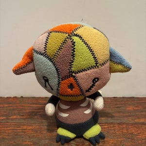 Op de afbeelding: Een handgemaakte pluche pop met een patchwork hoofd in verschillende kleuren, waaronder oranje, geel en blauw. De pop heeft zwarte gestikte details voor de ogen en mond, een bruine torso met een ribbenkastontwerp en een donkerblauwe short.