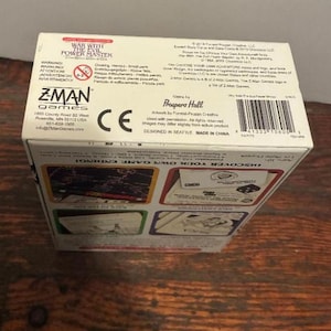 K&ouml;nnte beinhalten: Eine wei&szlig;e Pappschachtel f&uuml;r das Brettspiel "War With The Evil Power Master". Die Schachtel enth&auml;lt Text in mehreren Sprachen, das Z-Man Games-Logo und einen Barcode. Die Schachtel steht auf einer Holzoberfl&auml;che.