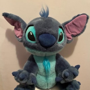 May include: A plush toy of the character Stitch with dark blue fur, a light blue chest and face patch, large ears with pink inner lining, black eyes with light blue accents, a dark blue nose, and a small tuft of blue hair on the top of the head.