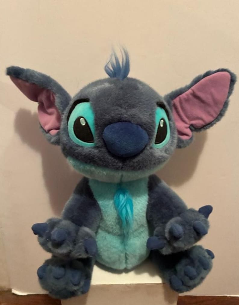 May include: A plush toy of the character Stitch with dark blue fur, a light blue chest and face patch, large ears with pink inner lining, black eyes with light blue accents, a dark blue nose, and a small tuft of blue hair on the top of the head.