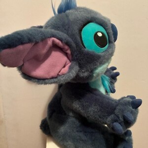 May include: A plush toy of the character Stitch, featuring dark blue fur, large ears with light purple inner lining, bright turquoise eye patches, and a light blue chest patch. The figure has small blue claws on its hands and a tuft of blue hair.