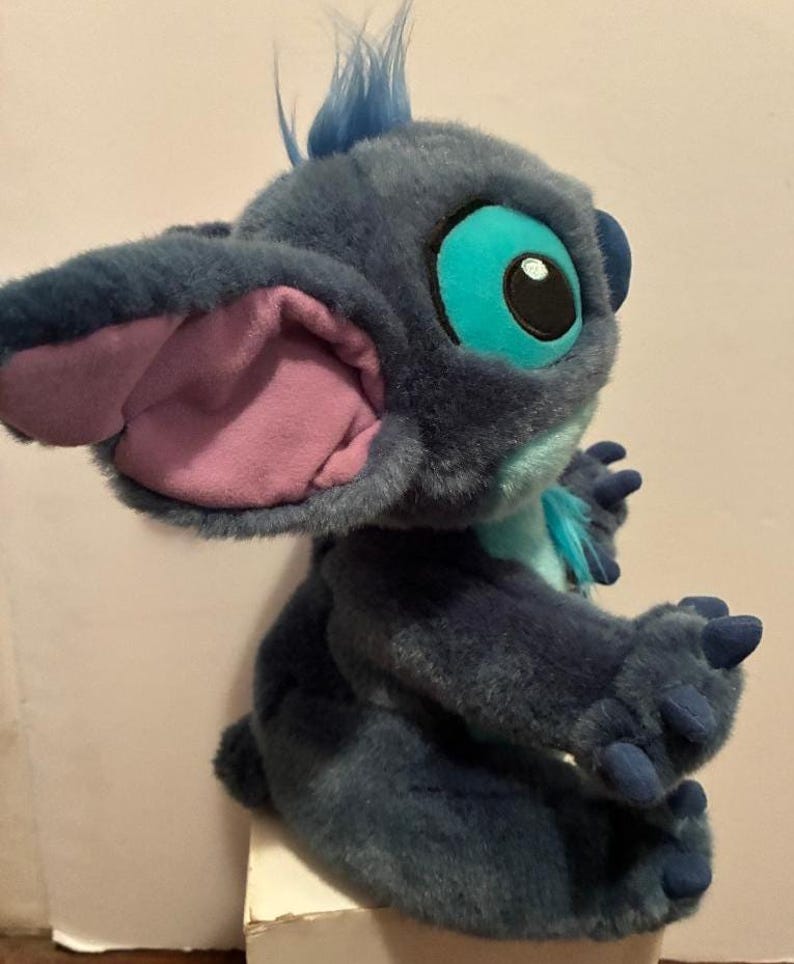 May include: A plush toy of the character Stitch, featuring dark blue fur, large ears with light purple inner lining, bright turquoise eye patches, and a light blue chest patch. The figure has small blue claws on its hands and a tuft of blue hair.