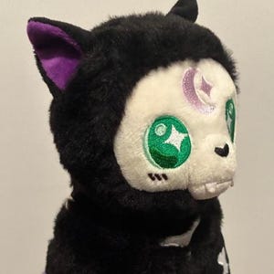May include: A plush black cat-like creature with a white face and green eyes. The ears have purple inner linings. A crescent moon and star are embroidered on the forehead. The creature is wearing a black outfit with a white skeleton design.