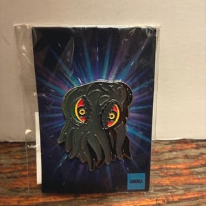 Op de afbeelding: Zwarte emaille pin met een gestileerd monsterontwerp met rode en gele ogen, tegen een donkerblauwe achtergrond met radiale lichteffecten. De pin zit in een transparante plastic hoes.