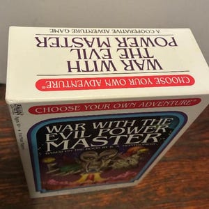 K&ouml;nnte beinhalten: Ein Brettspielkarton mit dem Titel "War With The Evil Power Master". Die Box ist wei&szlig; mit roten und blauen Akzenten. Der Titel des Spiels ist deutlich sichtbar, zusammen mit dem Satz "Choose Your Own Adventure". Die Box steht auf einer Holzoberfl&auml;che.