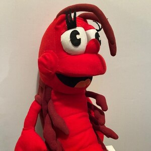 May include: A red plush lobster toy with large white and black eyes, a smiling mouth, and dark red antennae. The toy has a soft, textured appearance and is likely designed for children.