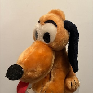 May include: A plush toy of a cartoon dog with orange fur, black ears, and a black nose. The dog has large, round eyes and a red tongue sticking out. The toy is sitting on a white surface.