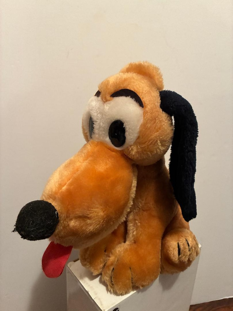 May include: A plush toy of a cartoon dog with orange fur, black ears, and a black nose. The dog has large, round eyes and a red tongue sticking out. The toy is sitting on a white surface.
