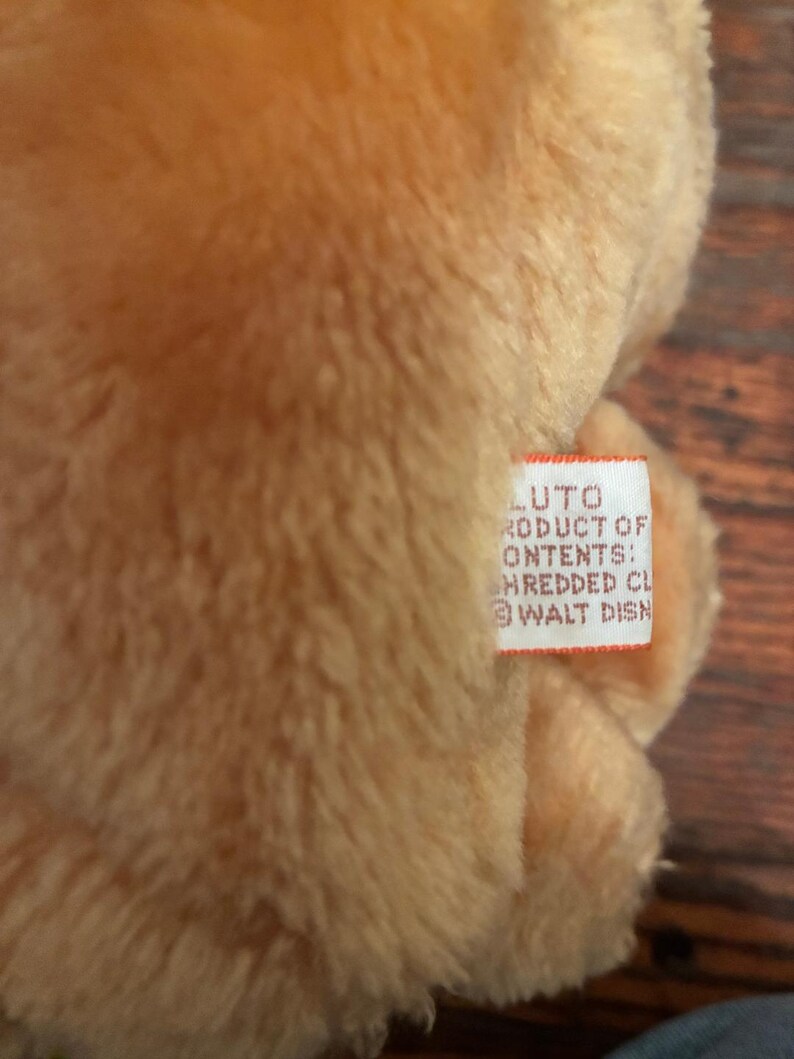May include: Close-up of a plush toy with a soft, textured, light brown fur. A small white tag is visible, with the text "LUTO PRODUCT OF CONTENTS: HREDDED CL @ WALT DISN".