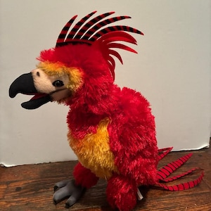 Op de afbeelding: Een pluche vogel met rode, gele en zwarte kleuren. De vogel heeft een rode kuif met zwarte strepen, een zwarte snavel en grijze poten. Het lichaam is voornamelijk rood met gele accenten.