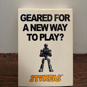 May include: A cream-colored box with the text "GEARED FOR A NEW WAY TO PLAY?" in black. Below the text is a black articulated action figure. The Stikfas logo is at the bottom in orange.