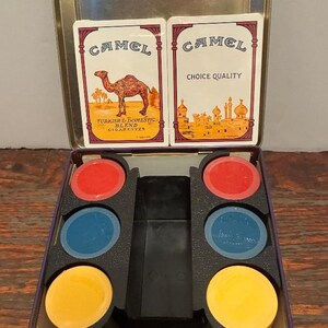 May include: Vintage Camel cigarette tin with playing cards and colorful game pieces. The tin features the Camel logo and text, with cards displaying a camel and Middle Eastern imagery. The game pieces are red, blue, and yellow.