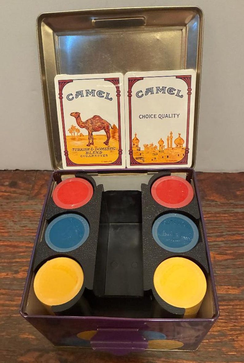 May include: Vintage Camel cigarette tin with playing cards and colorful game pieces. The tin features the Camel logo and text, with cards displaying a camel and Middle Eastern imagery. The game pieces are red, blue, and yellow.