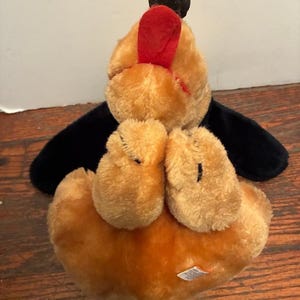 May include: A plush toy dog with a black head and wings, tan body, and red accents. The dog is lying on its back, with its paws and belly visible. The toy has a soft, fuzzy texture.