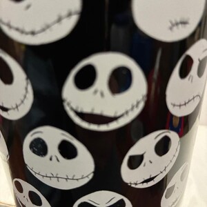 Puede incluir: Vaso negro con m&uacute;ltiples caras blancas de Jack Skellington. Las caras muestran diferentes expresiones, como sonrisas y ce&ntilde;os fruncidos. El dise&ntilde;o se repite alrededor del vaso, creando una est&eacute;tica espeluznante y divertida.