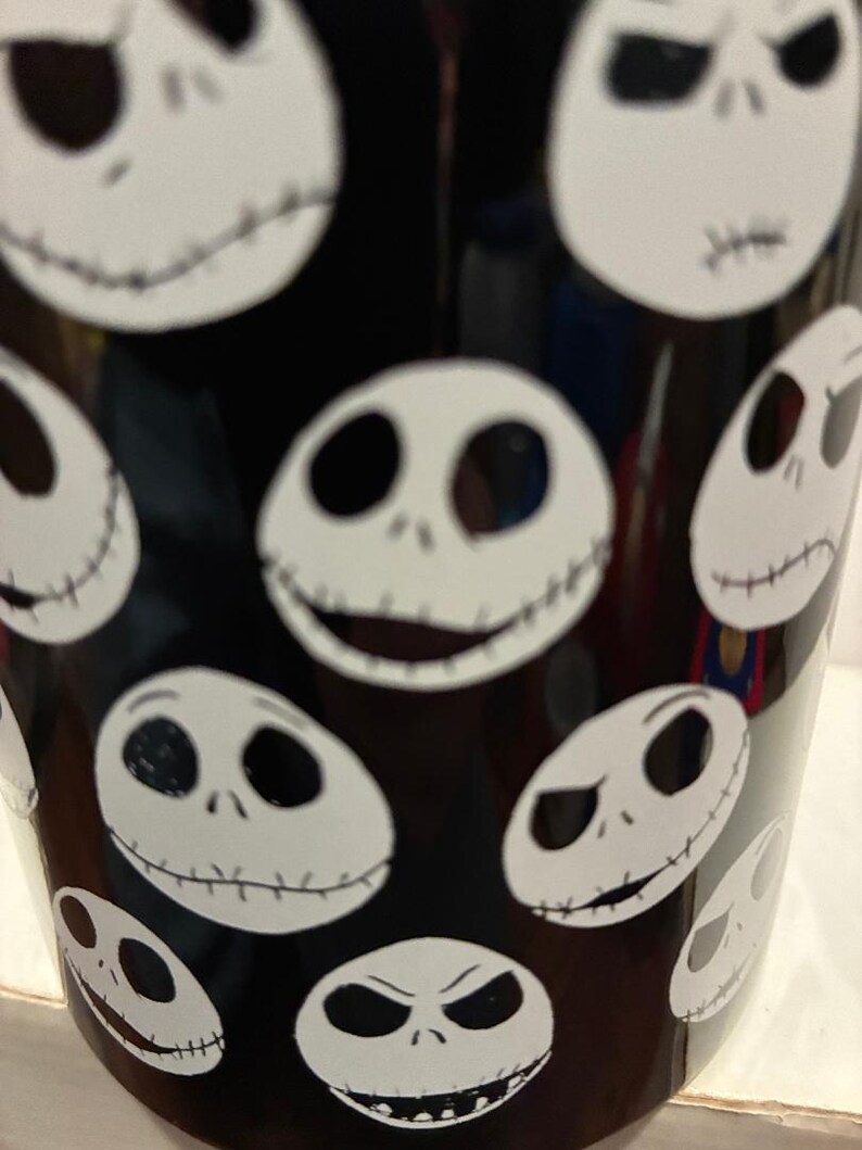 Puede incluir: Vaso negro con m&uacute;ltiples caras blancas de Jack Skellington. Las caras muestran diferentes expresiones, como sonrisas y ce&ntilde;os fruncidos. El dise&ntilde;o se repite alrededor del vaso, creando una est&eacute;tica espeluznante y divertida.