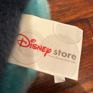 May include: A white fabric tag attached to a plush toy with soft blue and dark blue fur. The tag features the red Disney store logo and the website address www.disneystore.com printed in gray text, along with faint gray circular patterns.