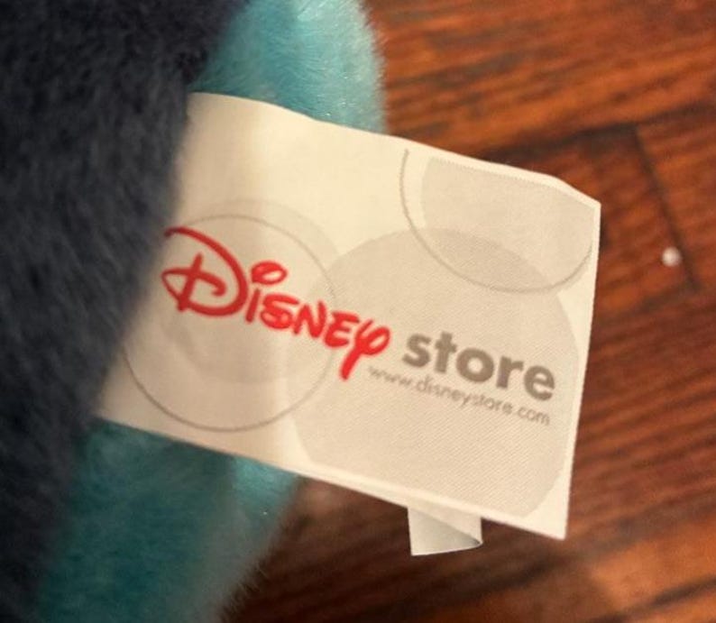 May include: A white fabric tag attached to a plush toy with soft blue and dark blue fur. The tag features the red Disney store logo and the website address www.disneystore.com printed in gray text, along with faint gray circular patterns.