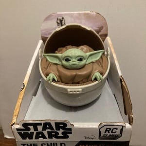May include: A Star Wars The Mandalorian The Child and Pram RC toy in its packaging. The toy is a green and brown figure of The Child, also known as Baby Yoda, sitting in a white pram. The packaging is white with blue and red accents and features the Star Wars logo, the Disney logo, and the text "The Child and Pram (RC)" and "Mandalorian".