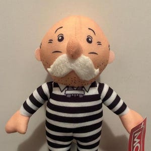 May include: A plush doll of the Monopoly Jail character, featuring a peach-colored head with a white mustache and black and white striped prison uniform. The doll has outstretched arms and a tag.