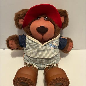 Op de afbeelding: Een bruine pluche beer met een rode baseballpet, een blauw-wit gestreept outfit en bruine laarzen. De beer heeft een bruine neus en zit rechtop. Het woord "Friskies" staat op de outfit.