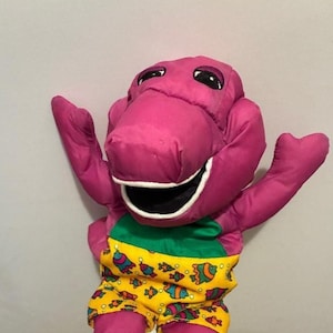 May include: A plush purple dinosaur toy with a white-lined mouth and black eyes. The dinosaur is wearing yellow shorts with a fish pattern and a green waistband. Its arms are outstretched, and it appears to be smiling.