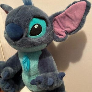 May include: A plush toy character with dark blue fur, a light blue chest patch, and a tuft of blue hair on its head. It features large, pointed ears with pink inner lining, a dark blue nose, and large eyes with light blue and black accents.