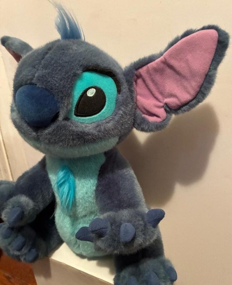 May include: A plush toy character with dark blue fur, a light blue chest patch, and a tuft of blue hair on its head. It features large, pointed ears with pink inner lining, a dark blue nose, and large eyes with light blue and black accents.
