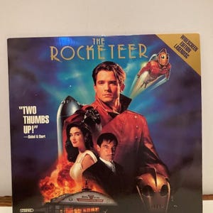 Peut inclure: Une jaquette de disque laser pour le film "The Rocketeer" avec un fond de ciel bleu, une fusée, un homme en veste rouge, une femme en robe blanche et un homme en costume. Le texte "Two Thumbs Up!" est écrit en blanc avec un contour noir. Le texte "Siskal & Ebert" est écrit en blanc plus petit en dessous. Le texte "Exciting Theatrical Trailer Included!" est écrit en blanc avec un contour noir en bas de la jaquette.