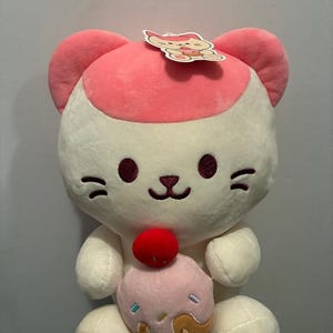 Könnte beinhalten: Ein weißes Plüschkatzenspielzeug mit rosa Akzenten an Ohren und Kopf. Die Katze hält einen rosa Cupcake mit einer roten Kirsche oben drauf. Ein kleines Etikett mit einer Katzenillustration ist am Kopf befestigt.