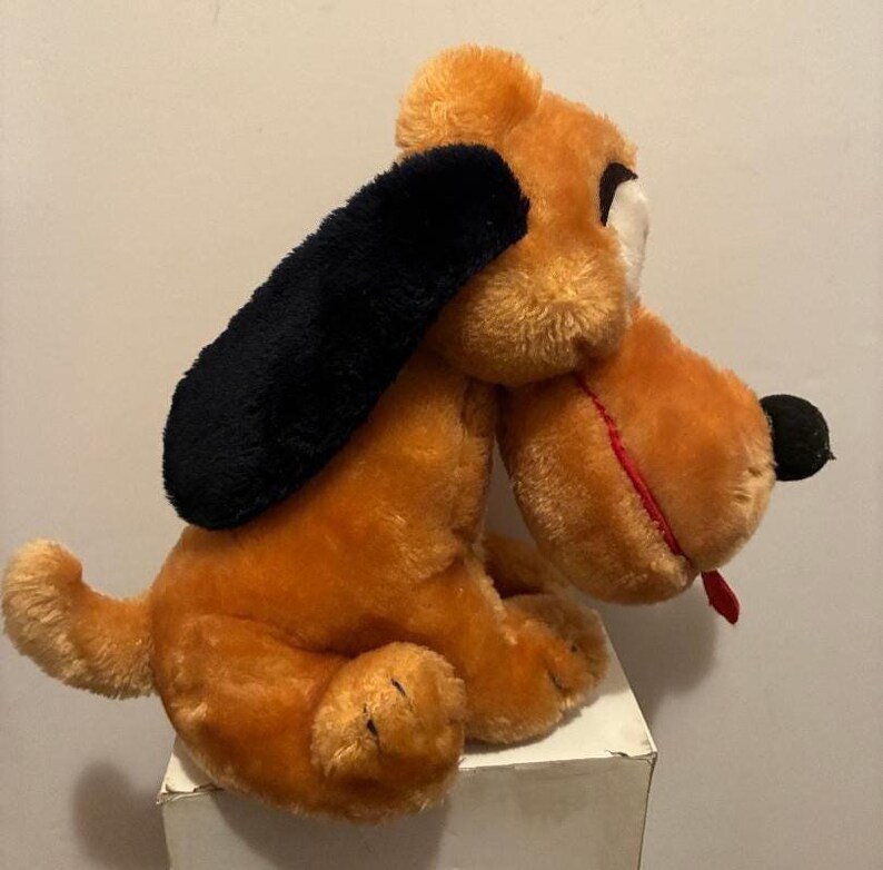 May include: A plush dog toy in shades of orange and brown, with a black ear and a red tongue. The dog is sitting on a white surface. The toy has a classic, vintage appearance.