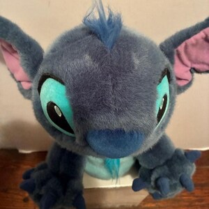 May include: A plush toy character with dark blue fur, large pointed ears with pink inner lining, and a tuft of blue hair on top. The character features large teal eye patches, black pupils, a dark blue nose, and three-fingered blue paws.