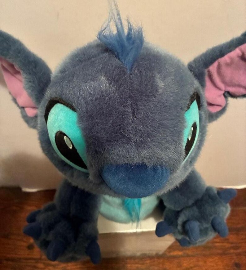May include: A plush toy character with dark blue fur, large pointed ears with pink inner lining, and a tuft of blue hair on top. The character features large teal eye patches, black pupils, a dark blue nose, and three-fingered blue paws.