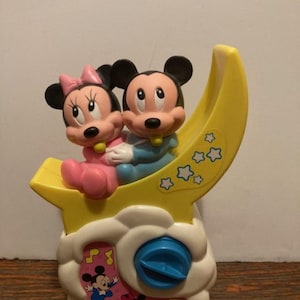 Könnte beinhalten: Vintage Disney-Musik-Nachtlicht mit Mickey- und Minnie-Mouse-Figuren. Die Figuren sind in Rosa und Blau gekleidet und sitzen auf einem gelben Halbmond. Die Basis ist weiß mit einem blauen Drehknopf und Noten.