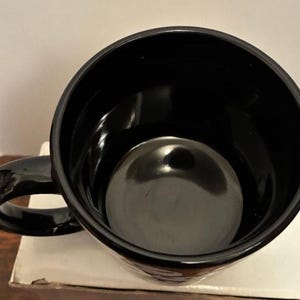 Puede incluir: Una taza de caf&eacute; negra brillante con un interior redondeado y un asa curva. La taza est&aacute; vac&iacute;a y parece estar hecha de cer&aacute;mica o un material similar. La taza est&aacute; sobre una superficie blanca.