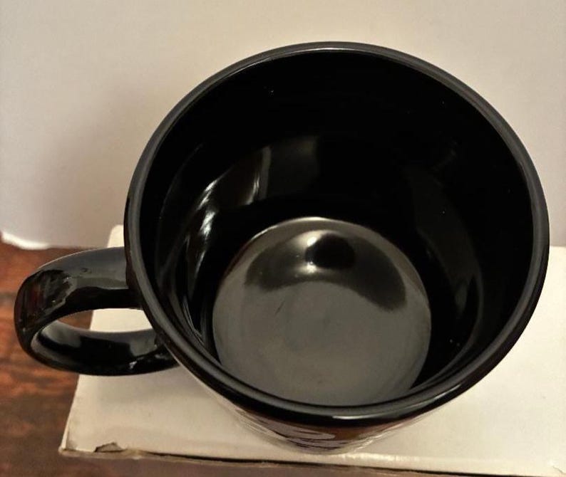 Puede incluir: Una taza de caf&eacute; negra brillante con un interior redondeado y un asa curva. La taza est&aacute; vac&iacute;a y parece estar hecha de cer&aacute;mica o un material similar. La taza est&aacute; sobre una superficie blanca.